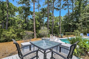A pool area with a glass table and chairs, surrounded by a wooden deck and dense greenery. - Huge Home, includes Bikes, Kayaks, Private Pool, Game Room, Dock... (Hilton Head Island)