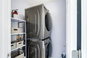A laundry room with a stacked washer and dryer, shelves with baskets, and a fire extinguisher. - Sunset Rentals | Starboard Tack 40 (Hilton Head Island)