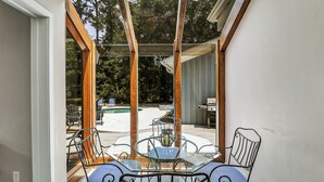 Un patio con paredes de vidrio, una mesa y sillas, una parrilla y una piscina al fondo.