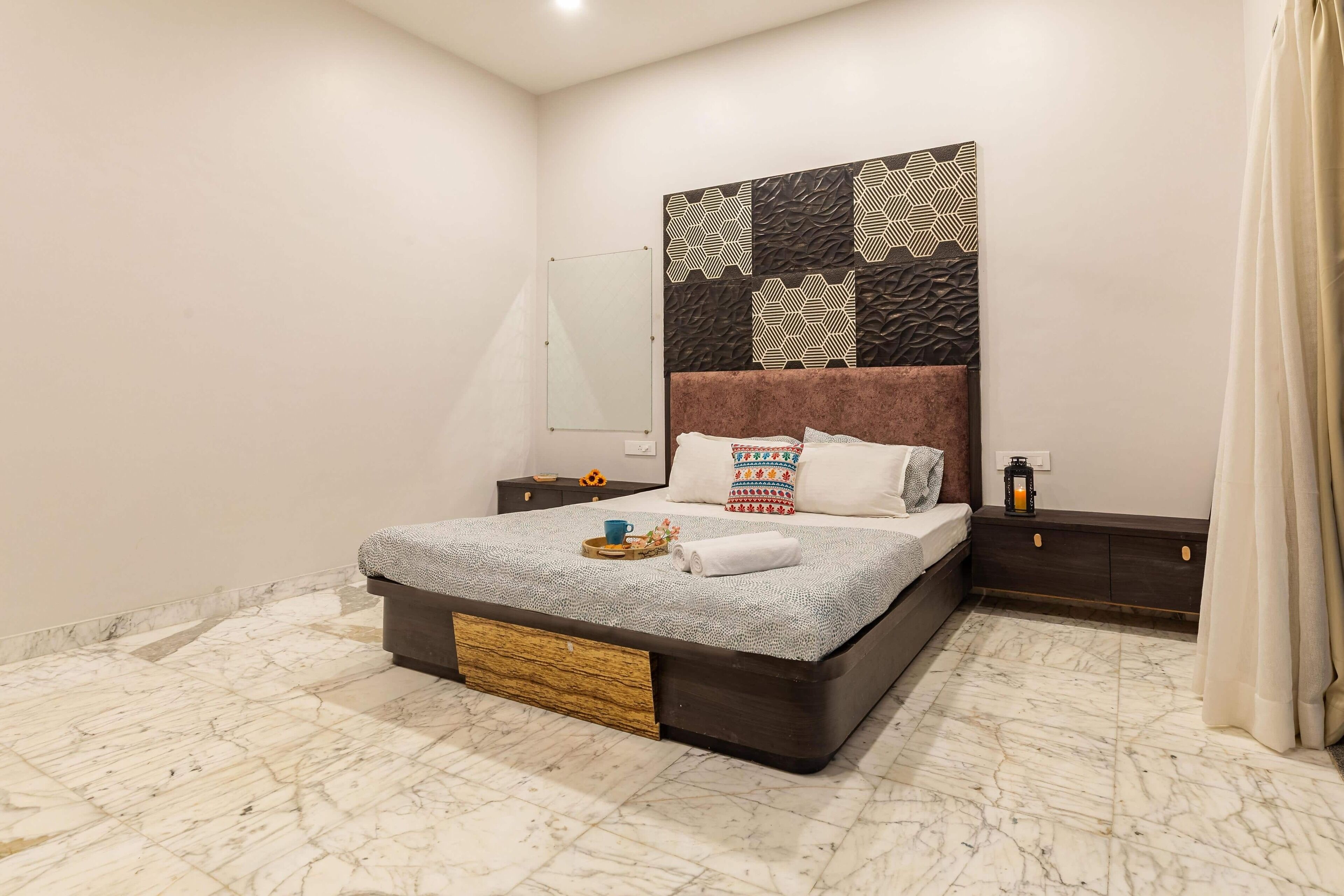 Un dormitorio moderno con suelo de mármol, cama con cubrecama estampado, cabecero decorativo y mesita de noche con un farol.