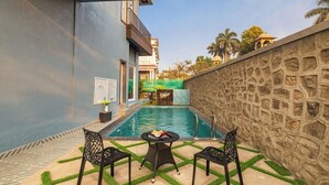 Une piscine avec deux chaises et une table, entourée d’un mur de pierre et d’un bâtiment bleu.
