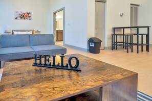 Un salon moderne avec un canapé, une table basse ornée des lettres formant le mot «BONJOUR», une poubelle et une table avec des chaises.