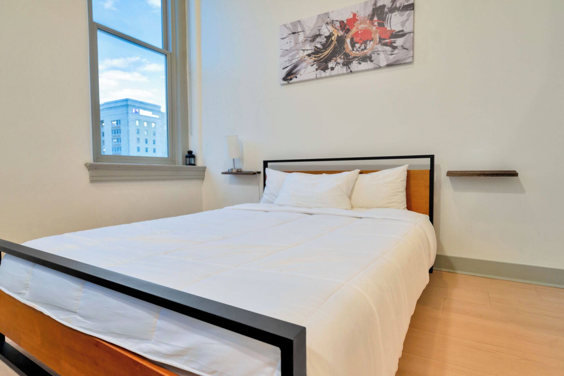 Ein ordentlich bezogenes Bett mit weißen Leinen in einem Zimmer mit großem Fenster, einem Wandbild und Holzboden.