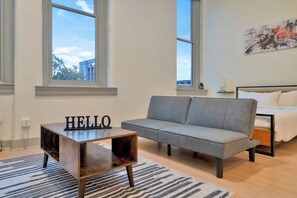 Ein modernes Schlafzimmer mit grauem Sofa, einem kleinen Holztisch mit der Aufschrift 'HELLO', einem Bett mit weißer Bettwäsche und einem großen Fenster mit Blick auf Gebäude.