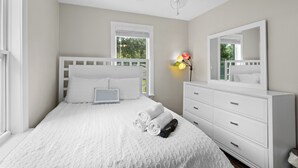 A bedroom with a white bed, a bedside table, a dresser, a ceiling fan, and a window with blinds. - Escape to this bright, pet-friendly cottage just minutes from Perdido Key's sugar-white sands! Two queen bedrooms sleep 4, plus a full kitchen, in-unit laundry, screened front porch & fenced backyard. Free driveway parking for 4! (Pensacola)