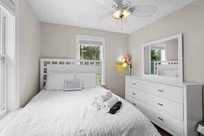 A bedroom with a white bed, a bedside table, a dresser, a ceiling fan, and a window with blinds. - Escape to this bright, pet-friendly cottage just minutes from Perdido Key's sugar-white sands! Two queen bedrooms sleep 4, plus a full kitchen, in-unit laundry, screened front porch & fenced backyard. Free driveway parking for 4! (Pensacola)