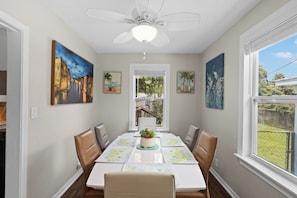 A dining area with a table set for four, surrounded by chairs, with artwork on the walls and a ceiling fan above. - Escape to this bright, pet-friendly cottage just minutes from Perdido Key's sugar-white sands! Two queen bedrooms sleep 4, plus a full kitchen, in-unit laundry, screened front porch & fenced backyard. Free driveway parking for 4! (Pensacola)