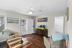 Un salon avec un ventilateur de plafond, un téléviseur posé sur un meuble en bois et deux fauteuils.