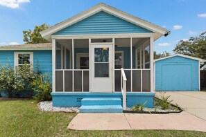 Une maison de plain-pied à l’extérieur bleu, avec un porche à l’avant doté d’une porte blanche et un petit jardin devant, avec un chemin en béton et quelques plantes.