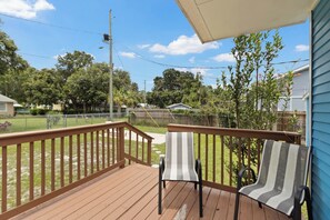 Une terrasse en bois avec deux chaises, une vue dégagée sur un jardin, et une maison bleue en arrière-plan.