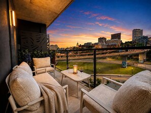 Ein Balkon mit Sitzgelegenheit und Tisch, mit Blick auf eine Skyline und eine Brücke bei Sonnenuntergang.
