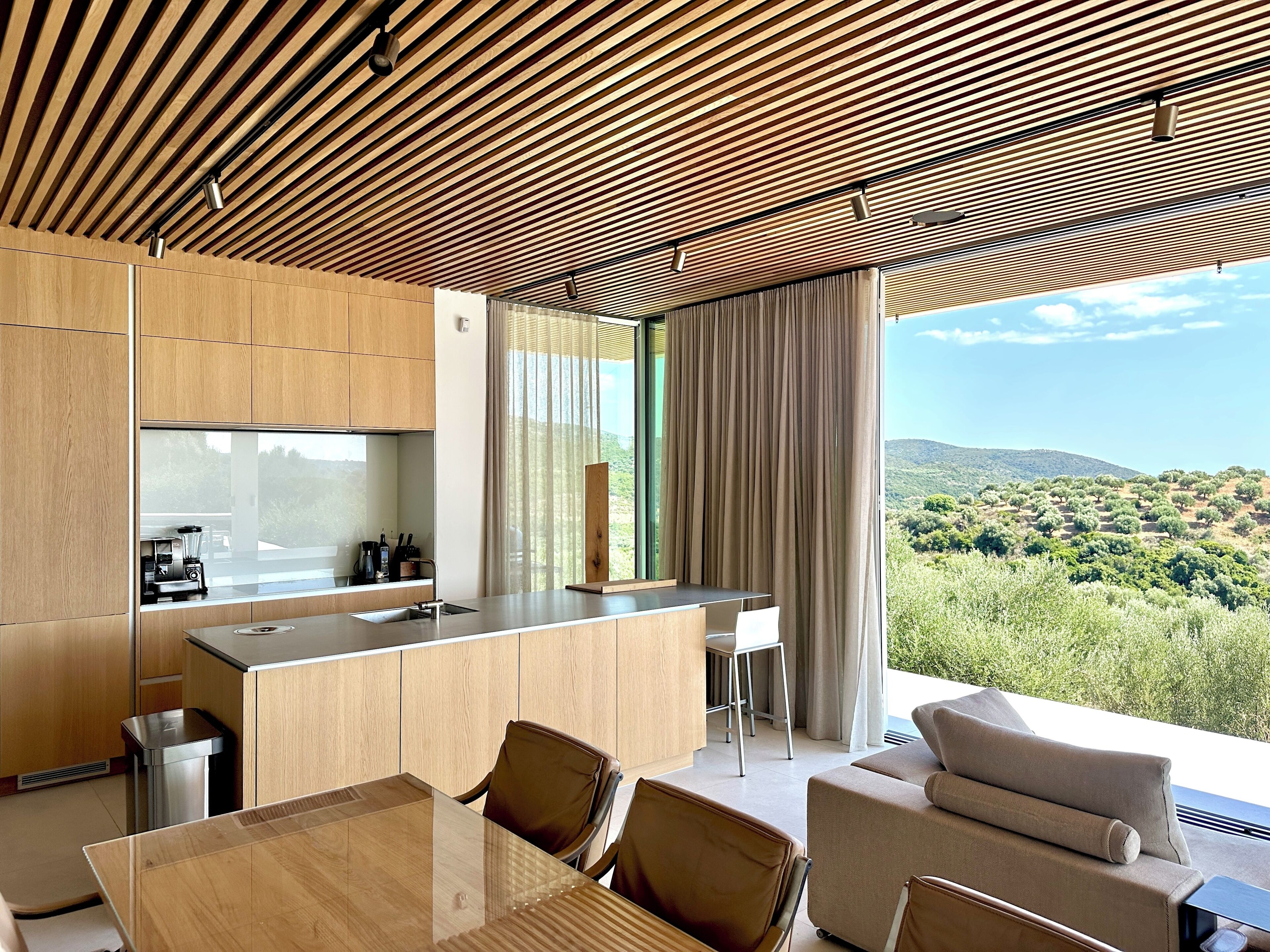 Une cuisine moderne dotée d’un îlot central, d’un coin repas avec des chaises et d’une grande fenêtre offrant une vue sur un paysage vallonné.