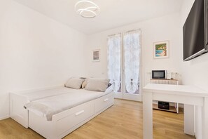 Ein schlicht eingerichtetes Schlafzimmer mit Bett, Schreibtisch, Fernseher und einem Fenster mit Vorhängen.
