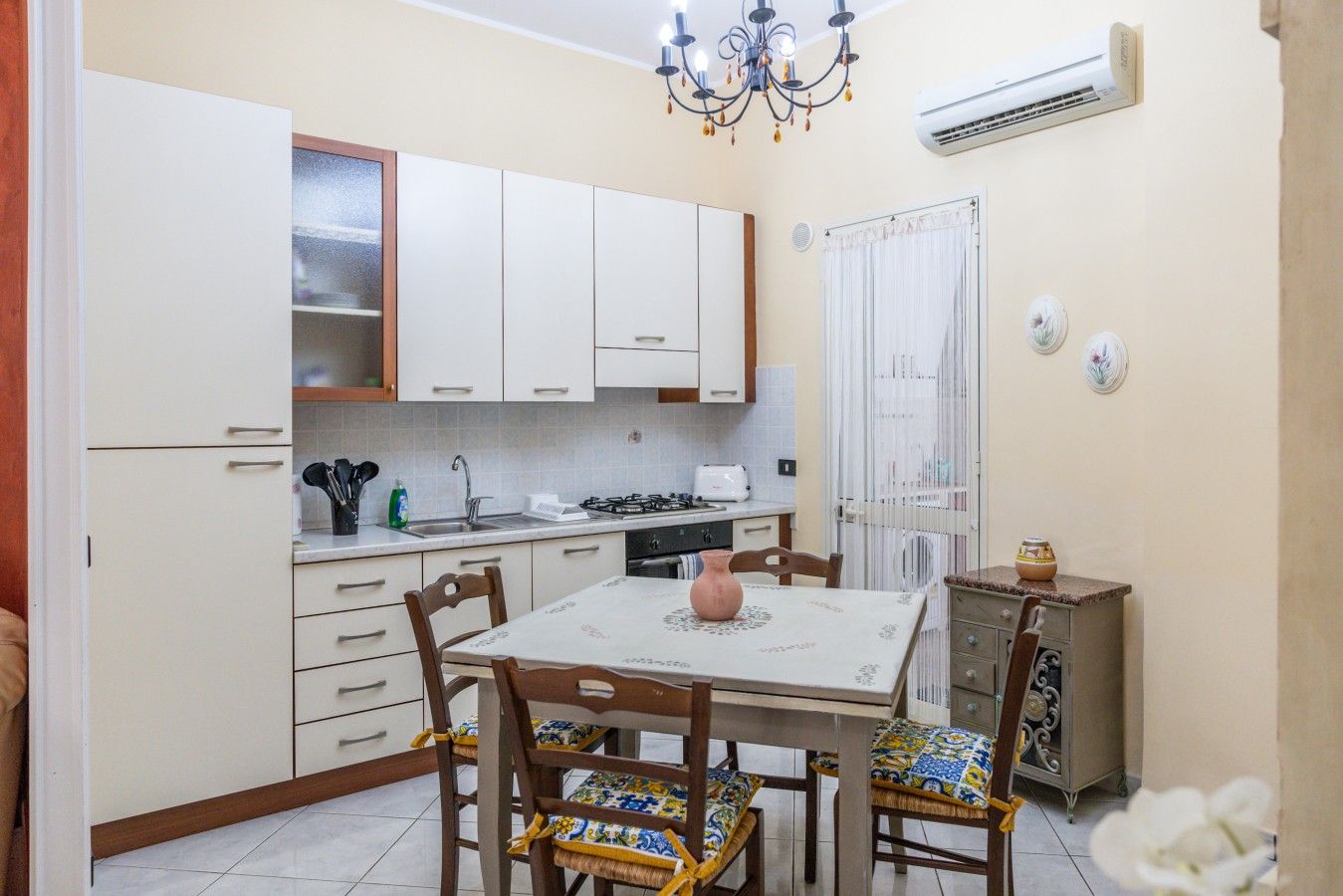 A compact kitchen with white cabinets, a dining table with chairs, and a small cabinet with decorative items.