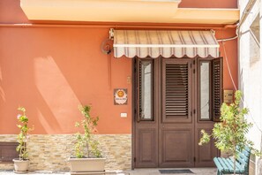 A building facade with a brown door, a striped awning, and two potted plants. - Casa del Sole - Menfi (Menfi)