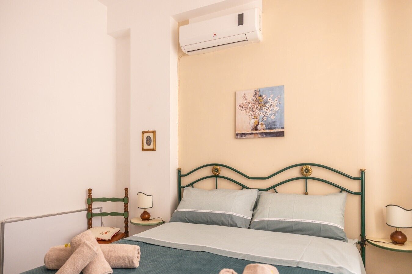 A bedroom with a bed, a nightstand, a lamp, a wall-mounted air conditioner, and a painting.