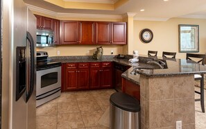 A kitchen with wooden cabinets, a stainless steel refrigerator, a built-in oven, a granite countertop, and a sink. - Phoenix West 2702 3 Bedroom Condo (Orange Beach)
