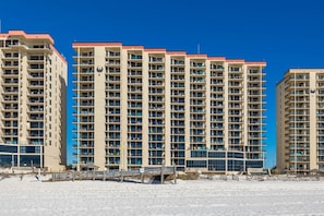 Un hotel moderno de gran altura con varios balcones, una playa arenosa al frente y un cielo azul despejado.