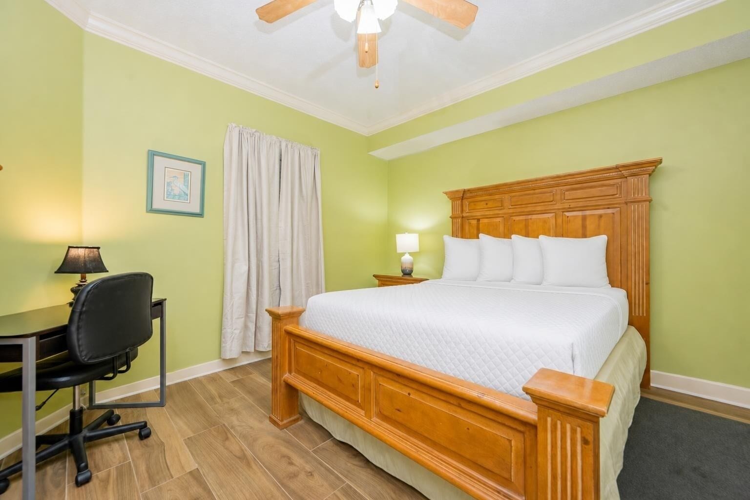 A hotel room with a wooden bed, a desk with a chair, a ceiling fan, and a window with curtains.
