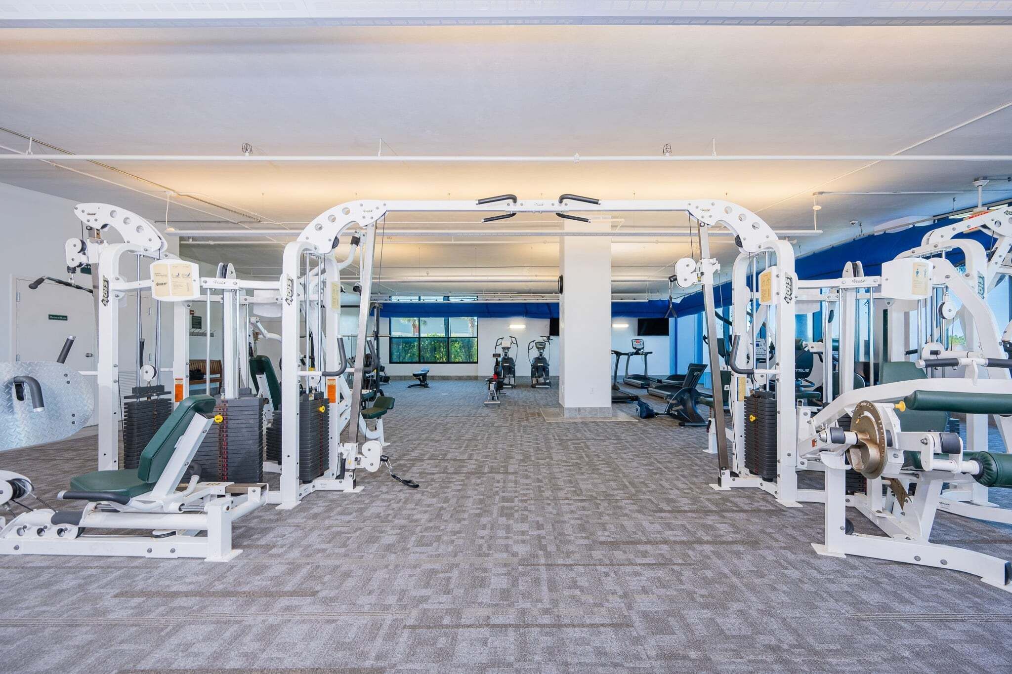 Un gimnasio con diversas máquinas de ejercicio, un espejo grande y vistas al exterior a través de amplias ventanas.