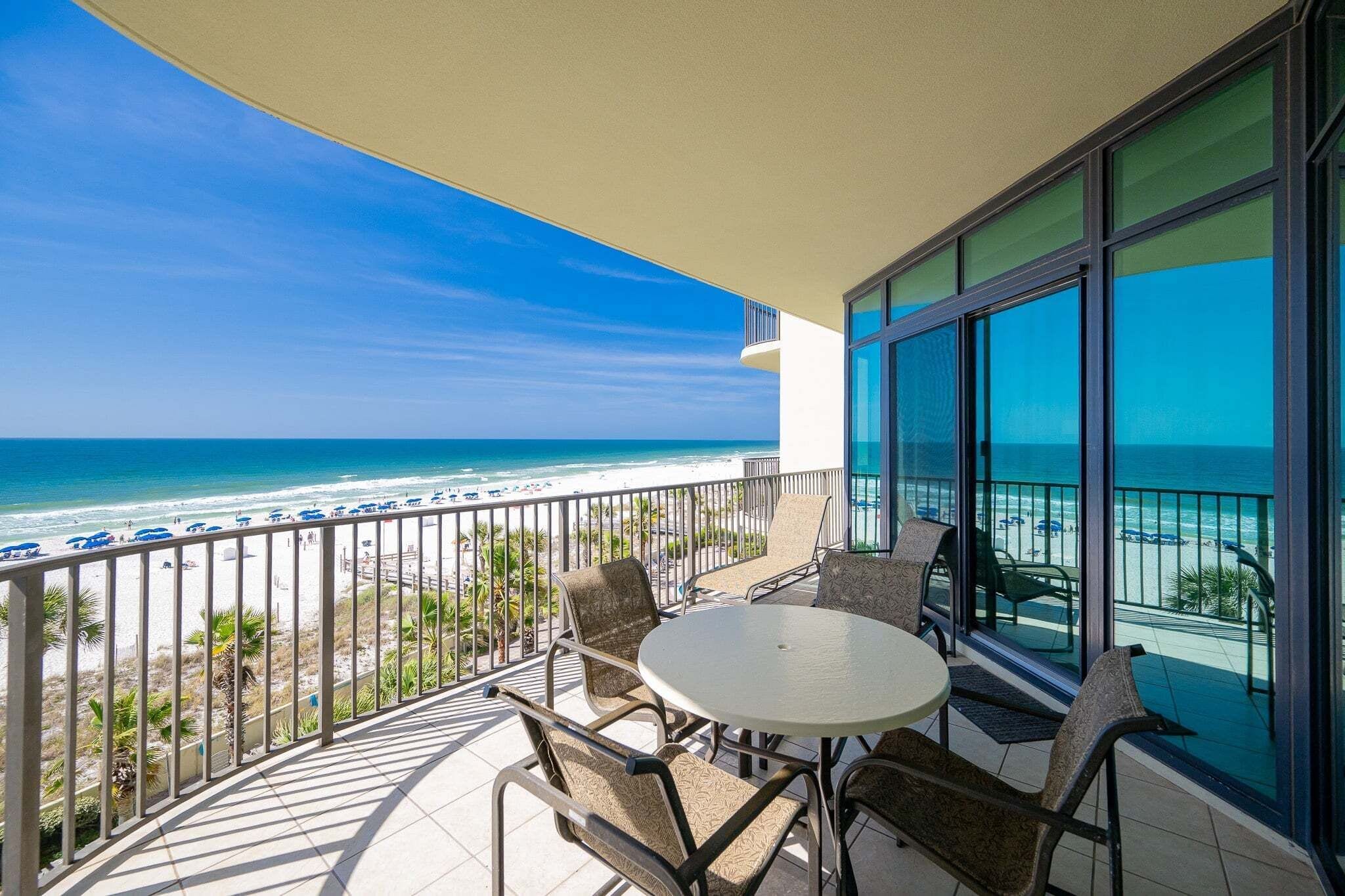 A balcony with a table and chairs, overlooking a beach with umbrellas and the ocean.