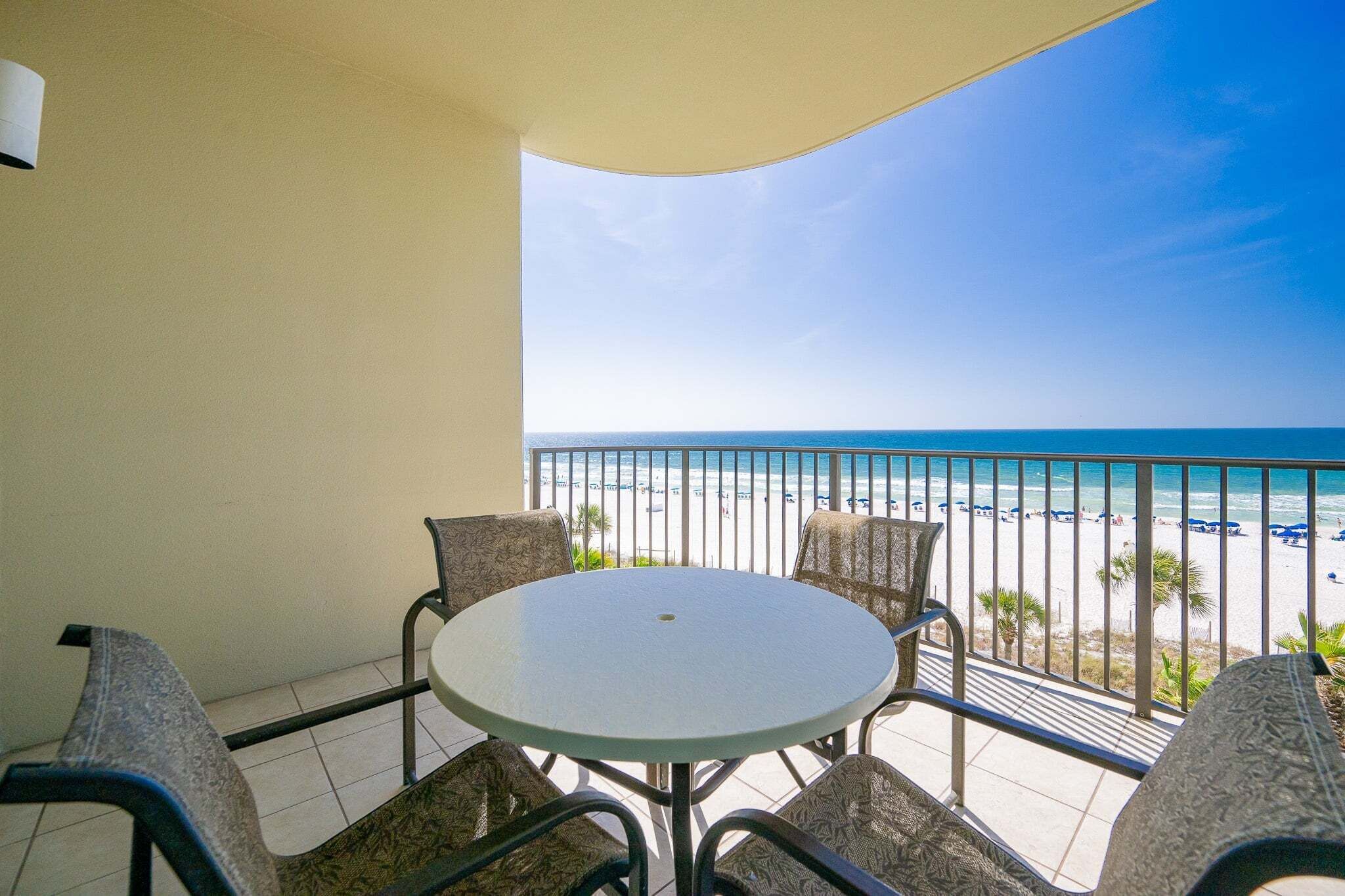 Ein Balkon mit einem runden Tisch und vier Stühlen, der Blick auf einen Strand unter wolkenlosem Himmel geht.
