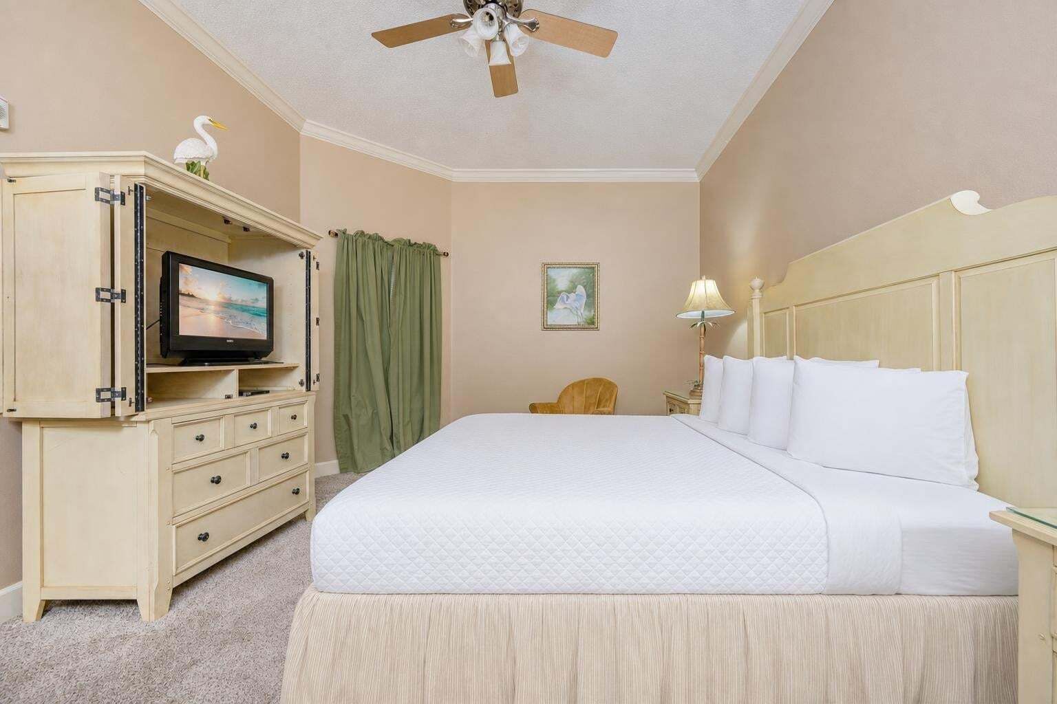 Habitación de hotel con una cama, un televisor sobre un mueble, un ventilador de techo, una silla y un cuadro en la pared.