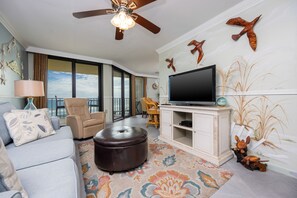 A living room with a sofa, ottoman, television, and decorative wall art. - Phoenix Iii 3055 1 Bedroom Condo (Orange Beach)