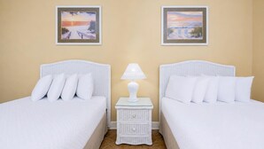 A hotel room with two beds, white bedding, a small white nightstand with a lamp, and two framed pictures on the wall. - Phoenix West 2006 3 Bedroom Condo (Orange Beach)