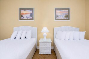 Ein Hotelzimmer mit zwei Betten, weißer Bettwäsche, einem kleinen weißen Nachttisch mit Lampe und zwei gerahmten Bildern an der Wand.