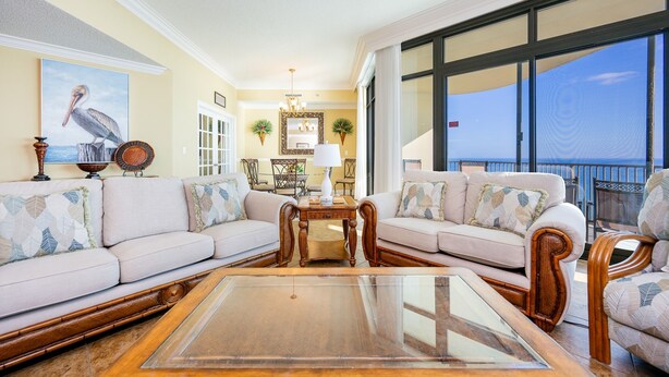 A spacious living room with a large glass coffee table, two sofas with patterned cushions, a dining area with a chandelier, and a view of the ocean through large windows. - Phoenix West 2006 3 Bedroom Condo (Orange Beach)
