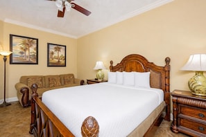 A hotel room with a large bed, a wooden headboard, a nightstand with a lamp, a sofa, and two wall-mounted paintings. - Phoenix West 2006 3 Bedroom Condo (Orange Beach)