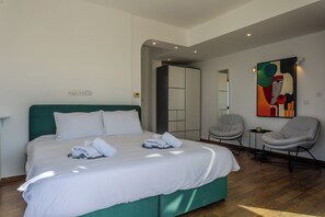 Moderni hotellihuone, jossa on suuri sänky, vihreäverhoiltu pääty, oleskelualue, jossa on kaksi tuolia, sekä värikäs abstrakti maalaus seinällä.