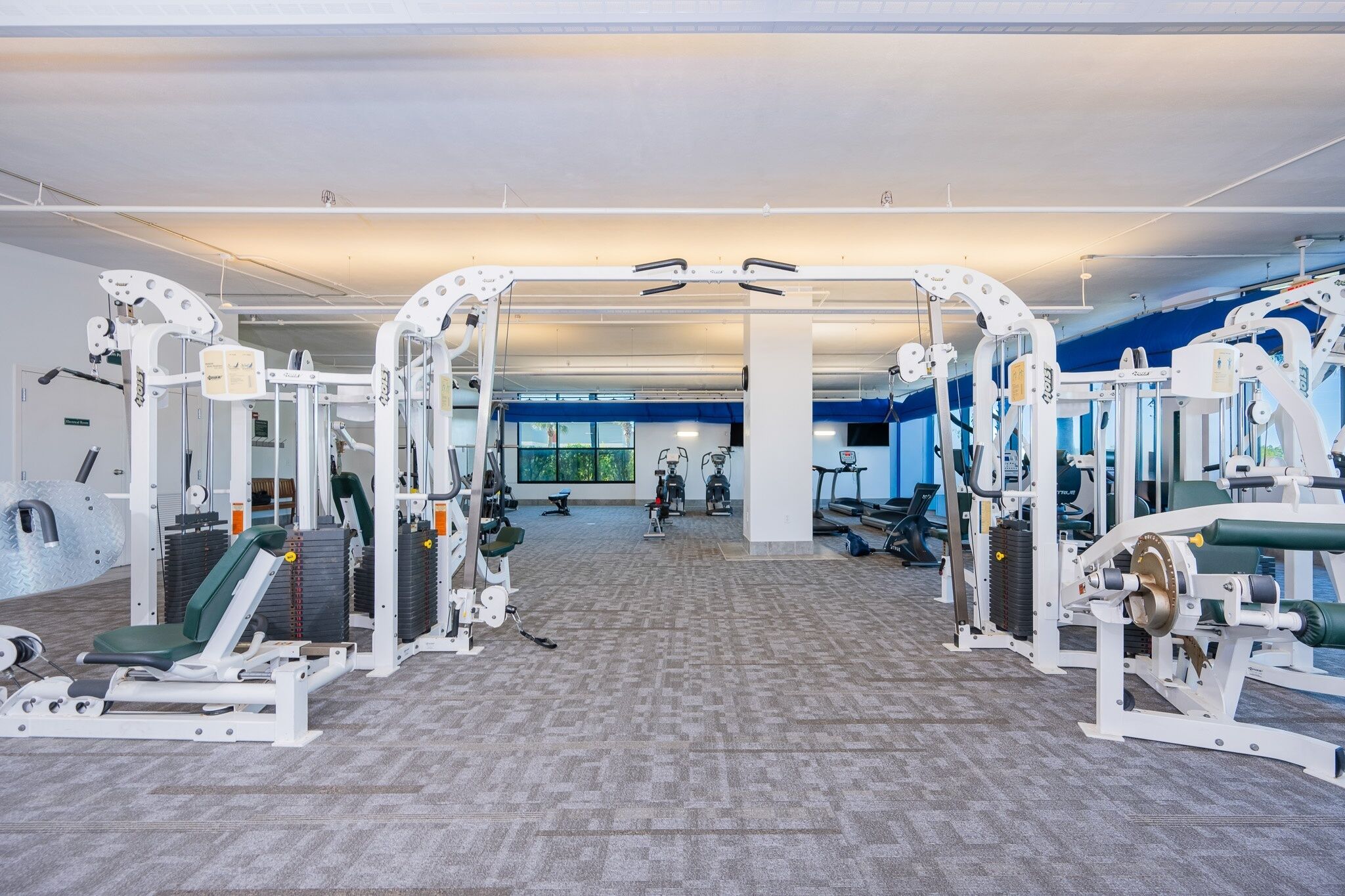 Un gimnasio con diversas máquinas de ejercicio, un amplio ventanal y suelo cubierto de alfombra.
