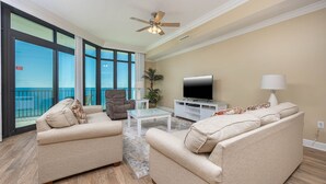 A living room with two beige sofas, a glass coffee table, a TV, and a ceiling fan. - Phoenix West 1006 3 Bedroom Condo (Orange Beach)