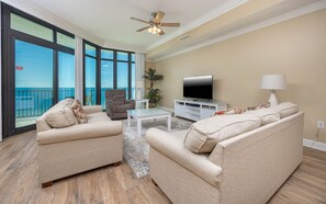 Ein Wohnzimmer mit zwei beigefarbenen Sofas, einem Glastisch, einem Fernseher und einem Deckenventilator.