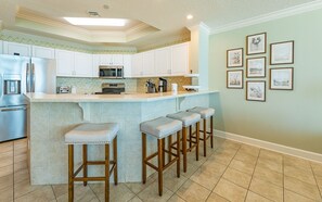 A kitchen with white cabinets, a built-in microwave, a stainless steel refrigerator, and a bar stool area. - Phoenix West 602 3 Bedroom Condo (Orange Beach)