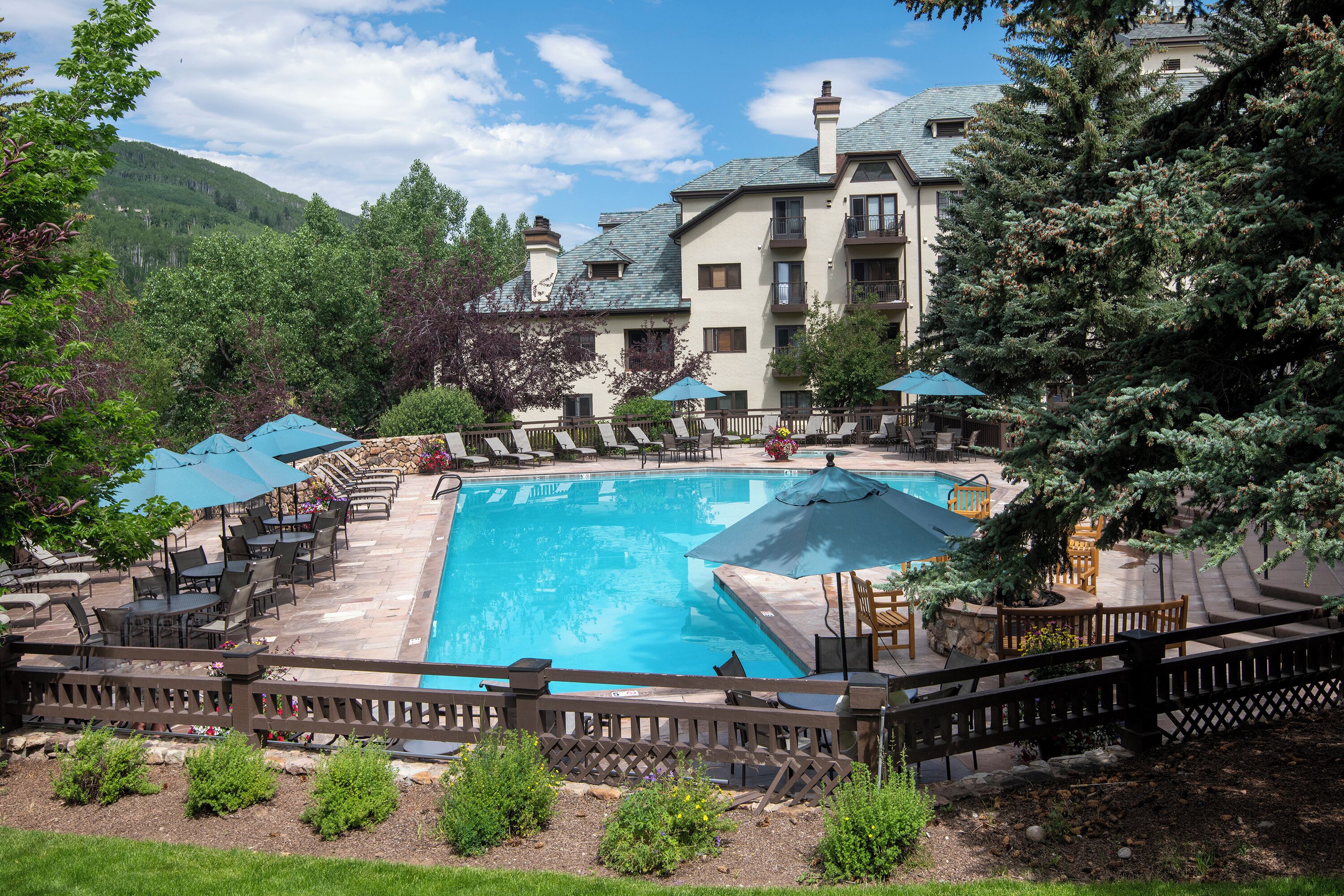 A swimming pool surrounded by lounge chairs and umbrellas, with a stone wall and a wooden fence.