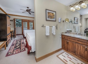 A hotel room with a bed, a desk, a chair, a bathroom with a sink and mirror, and a ceiling fan.