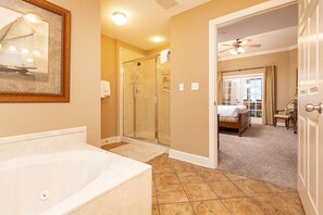A bathroom with a bathtub, a glass shower enclosure, and a door leading to a bedroom with a bed and a chair. - Phoenix West 2603 3 Bedroom Condo (Orange Beach)