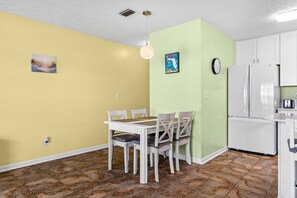 A compact kitchen with white cabinets, a white refrigerator, a dining table with chairs, and a hanging pendant light. - Wendwood F2 | Walk to the Beach • Community Pool • Sleeps 5 (Panama City Beach)