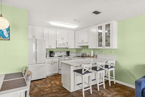 A compact kitchen with white cabinets, a microwave, a stove, and a sink. There are bar stools and a small dining table. - Wendwood F2 | Walk to the Beach • Community Pool • Sleeps 5 (Panama City Beach)