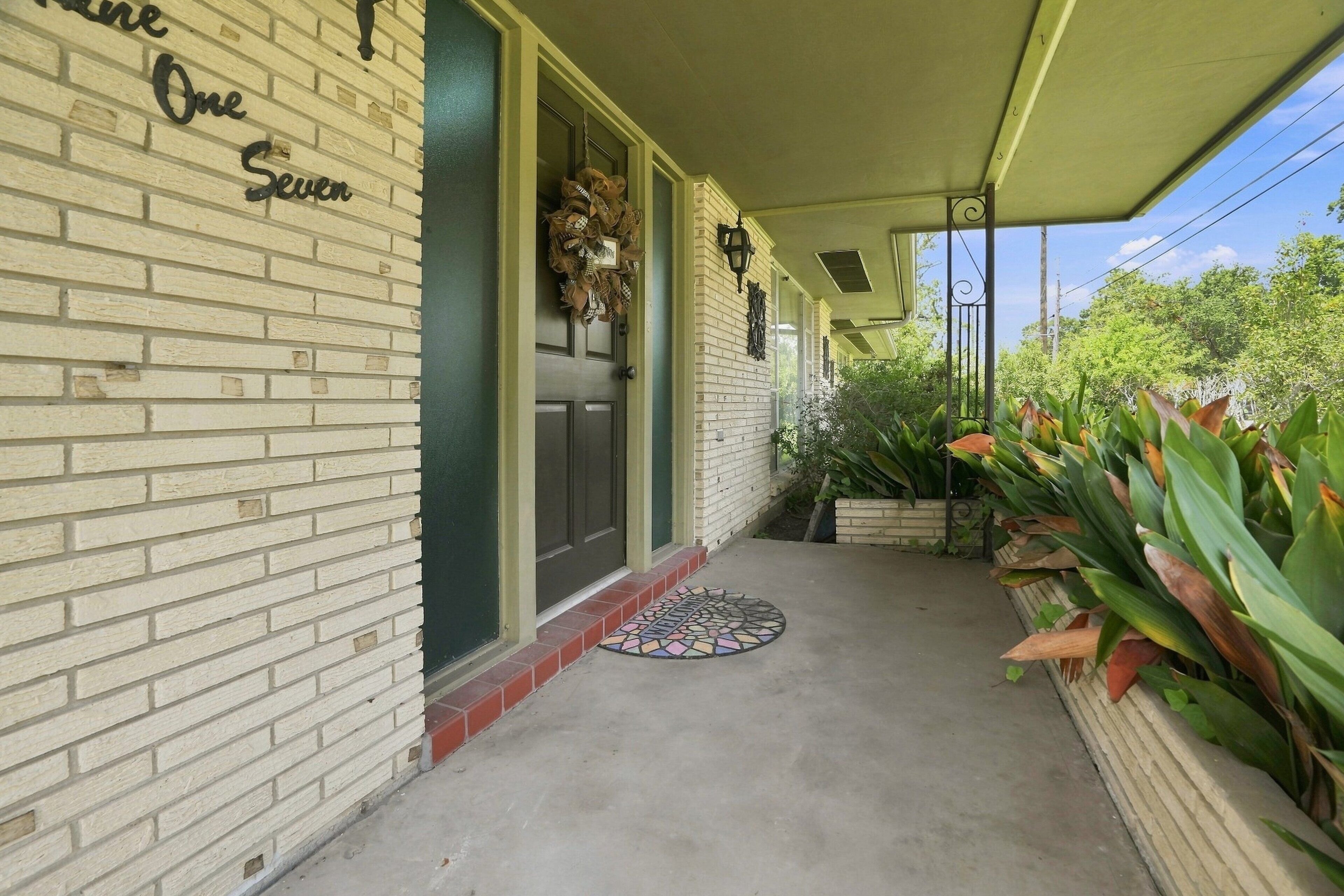A covered porch with a brick wall, a door with a wreath, and a tiled entry mat.