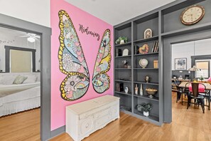 Ein Zimmer mit rosaroter Wand, einer weißen Kommode, einem Bücherregal und einer Essecke mit Tisch und Stühlen.