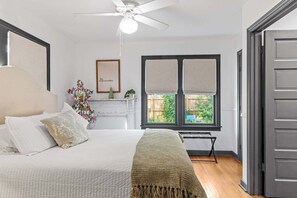 Ein Schlafzimmer mit Bett, Deckenventilator, Fenster mit Jalousien und Blick ins Freie.