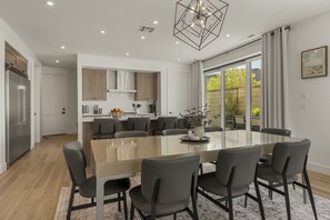 A modern kitchen with a central island, a dining area with a glass table, and a view of the outdoors. - Courtside Sunsets 5 Bedroom Townhouse (St. George)