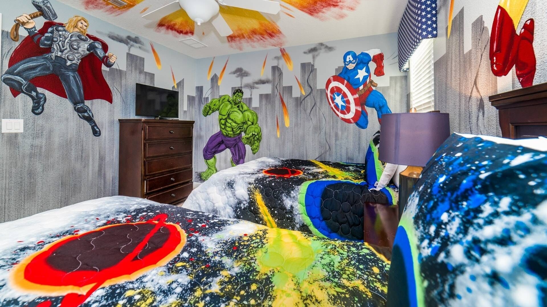 Une chambre avec un lit agrémenté d’une couvre-lit sur le thème des super-héros, une commode et un ventilateur de plafond.