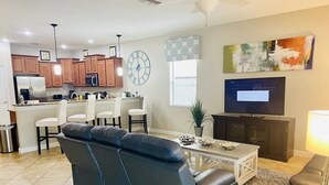 A modern living room with a sofa, a dining area, kitchen cabinets, a television, and a clock on the wall. - Wex1410 9 Bedroom Home (Davenport)