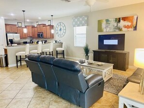 A modern living room with a sofa, a dining area, kitchen cabinets, a television, and a clock on the wall. - Wex1410 9 Bedroom Home (Davenport)