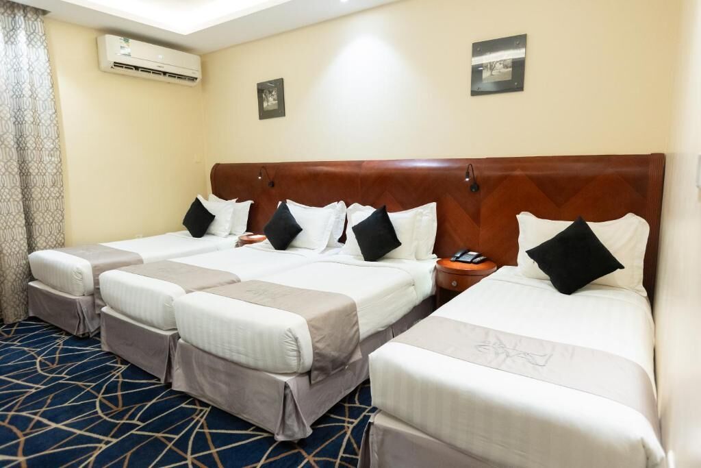 A hotel room with three beds, a patterned carpet, an air conditioning unit, and two framed pictures on the wall.